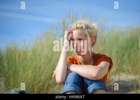 A thoughtful looking woman sitting on the sandy dunes surrounded by grass Stock Photo