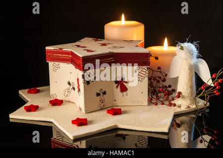 Christmas decoration, red and white star-shaped box with angels on, two lit candles, felted star, black background Stock Photo