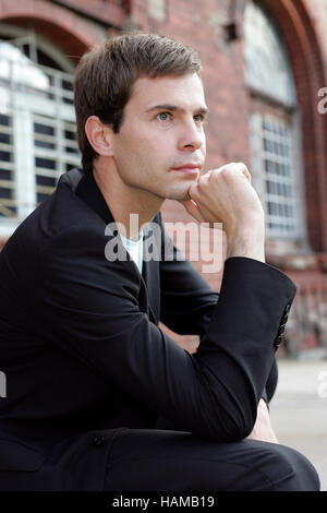 Portrait of a young man wearing a suit, sitting in front of an industrial building made of red bricks and thinking Stock Photo