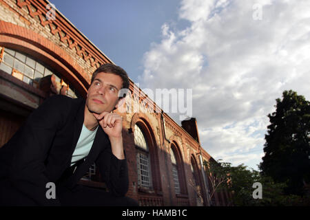 Portrait of a young man wearing a suit, sitting in front of an industrial building made of red bricks looking into the distance Stock Photo