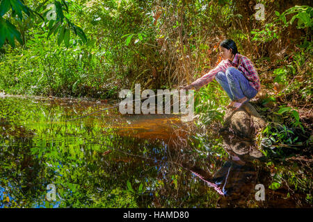 A young Khmer Cambodian woman squats beside a forest stream fishing for her family in Banteay Srei, Kingdom of Cambodia. Stock Photo