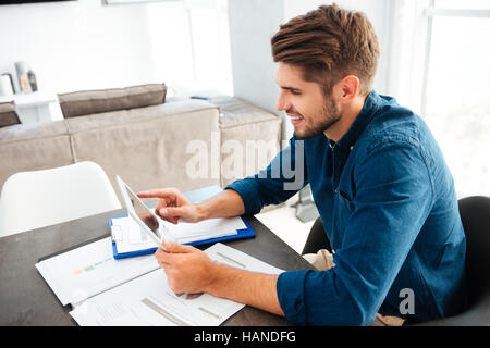 Photo of happy young man dressed in blue shirt sitting at home near documents and analyzing finances while holding tablet in hands and smiling. Lookin Stock Photo