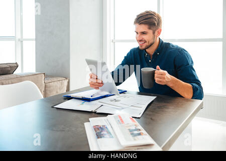 Young sman dressed in blue shirt holding tablet in hand and drinking coffee while sitting with documents and smiling. Stock Photo
