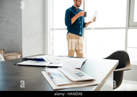 Picture of man's table with documents and newspapers. Young man standing near big window and drinking coffee while reading newspaper at the background Stock Photo