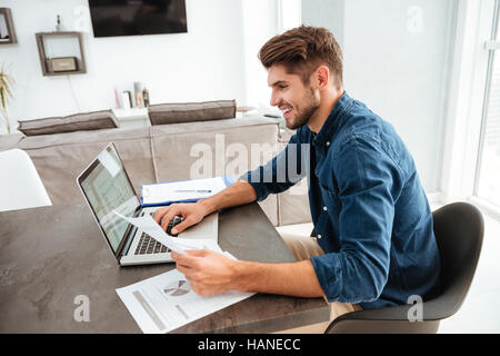 Photo of happy man using laptop and sitting at the table while looking at the papers. Stock Photo