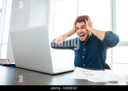 Photo of young shocked young man dressed in blue shirt while holding his head with hands and looking at laptop. Stock Photo