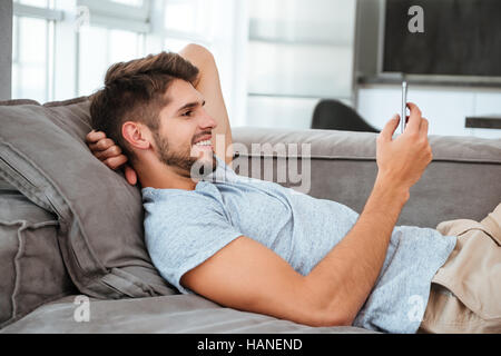 Picture of cheerful young man lies on sofa and looking on phone. Stock Photo