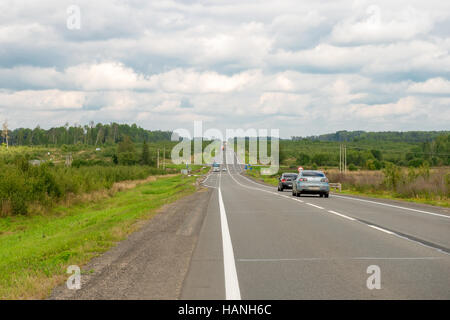 Asphalt road through the green fields and the cars traveling on it. Wooded mountains in the background. Summer day with white clouds. Stock Photo