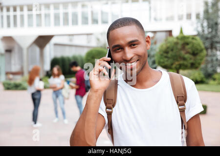 Happy african american young man student with backpack standing and talking on cell phone outdoors Stock Photo