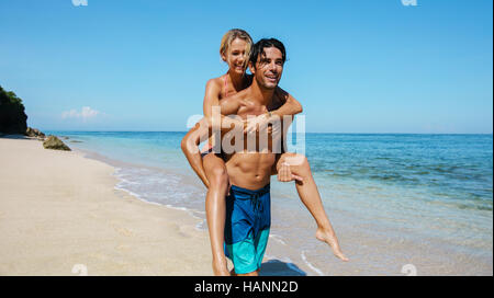 Portrait of handsome young man carrying girlfriend on his back and walking along the coast. Couple enjoying piggyback ride on the beach. Stock Photo