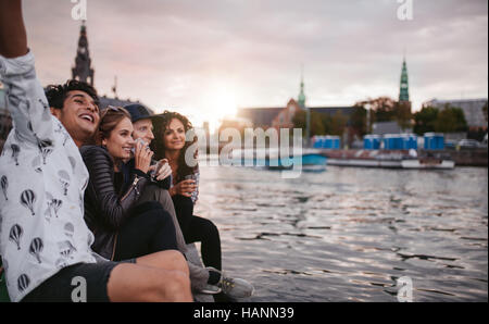Shot of group of people sitting outdoors on jetty and having fun. Friends hanging out on pier over lake in the city. Stock Photo