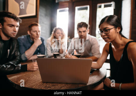 Group of friends sitting around cafe table and looking at laptop. Young men and women at coffee shop using laptop computer. Stock Photo