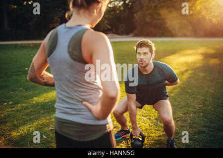 Female trainer guiding man while exercising with kettlebell in park. Personal trainer with man doing weight training in park. Stock Photo