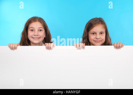 Two happy smiling beautiful young girl showing blank signboard or copyspace for slogan or text, isolated on blue background Stock Photo