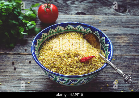 Raw bulgur wheat grains in colorful arabic bowl, fresh parsley, tomato and peppers for cooking. Wooden table background Stock Photo