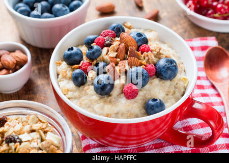 Oatmeal porridge with nuts, blueberries and raspberries in red bowl, close up view. Healthy breakfast, diet concept. Stock Photo