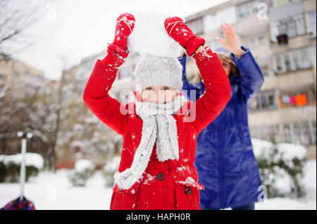 Mother with daughter cheerfully spend time in winter day. They are dressed in warm bright down-padded coats. Girl holds a big lump of snow on the head Stock Photo