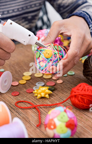 closeup of a young man making a handmade christmas ball using a hot glue gun with strings and buttons of different colors, and a pile of different hab Stock Photo