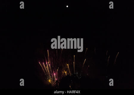 Golden fireworks with pink and yellow highlights amidst columns of smoke rising like rockets towards a bright full moon Stock Photo
