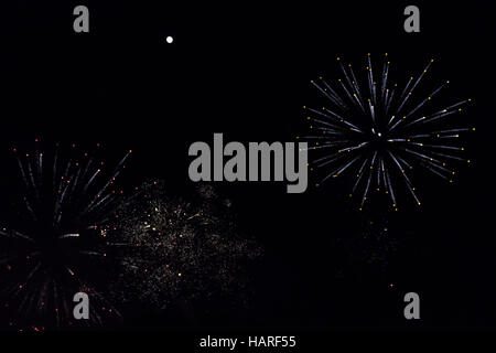 Colorful fireworks and white sparklers beneath a bright full moon nearing its apex in a dark rural sky Stock Photo