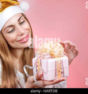 Portrait of a cute Santa girl wearing golden festive hat with gift box in hands isolated on pink background, beautiful Santa helper, happy Christmas Stock Photo