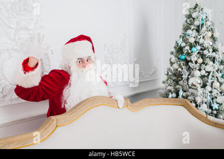 Santa Claus standing behind a Christmas tree and sofa looking at the camera Stock Photo