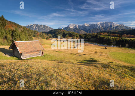 Haystack above Lake Gerold and the Karwendel mountains surrounded by colorful humpy meadows and autumn forest in afternoon sunlight, Bavaria, Germany Stock Photo