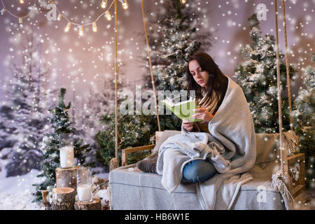 Romantic young woman reading a book while sitting on a swing and wrapped in a warm blanket in a snow-covered park with spruce trees, while snowing, co Stock Photo