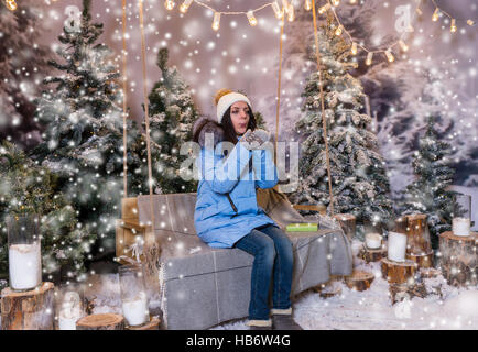 Female in blue down jacket blows snowflakes while sitting on a swing with a blanket under the flashlights in a snow-covered park with spruce trees, we Stock Photo