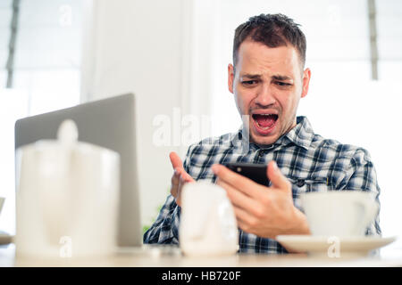 Bad news. Angry young man holding a mobile phone and grimacing at cafe Stock Photo