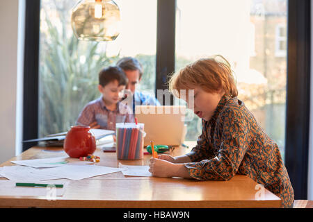 Boy colouring at dining table whilst father using laptop with brother Stock Photo