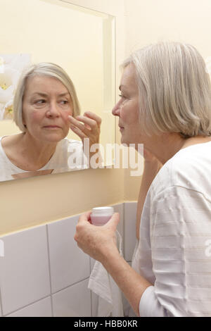 Elderly lady with a cream jar in hand standing in front of a mirror in a yellow bathroom and applying moisturizer to her face Stock Photo
