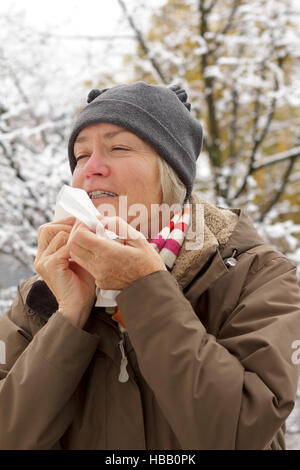 Senior woman with a red nose in winter coat, scarf and hat sneezing into a paper handkerchief in front of a tree with snow Stock Photo