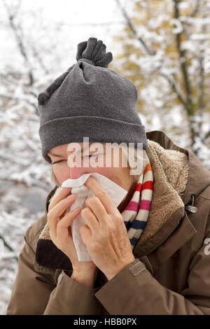 Senior woman with scarf and hat in a winter jacket blowing her nose into a paper tissue in front of a tree with snow Stock Photo