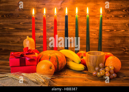 Kwanzaa holiday concept with traditional lit candles, gift box, pumpkins, ears of wheat, grapes, orange, banana, bowl and fruits on wood background Stock Photo