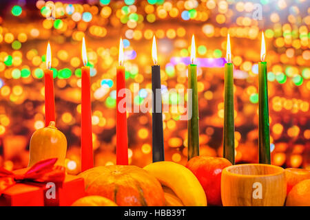 Kwanzaa celebration concept with seven candles red, black and green, gift box, pumpkin, bowl and fruit on light blur bokeh background, close up Stock Photo