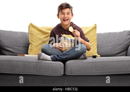 Cheerful boy sitting on a sofa and eating potato chips isolated on white background Stock Photo