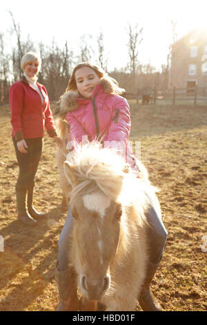 Young girl riding pony, mother watching from behind Stock Photo