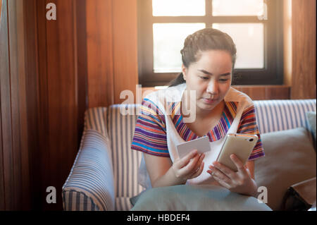 Trendy weekend lifestyle. Young woman using phone for on line shopping and another hand holding credit card while sitting on a couch. Shopping at home Stock Photo