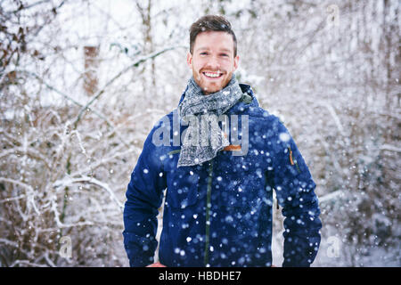 Young handsome man in warm coat smiling at camera standing in snowfall on background of snowy forest. Stock Photo