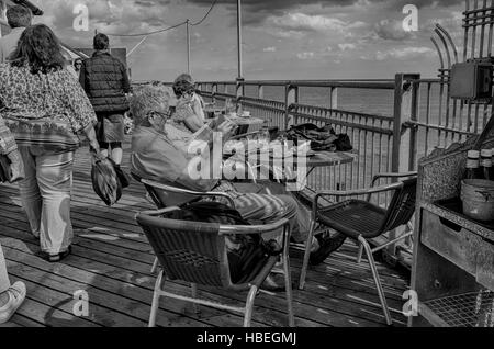 Southwold, England. Elderly people enjoying the sunny weather sitting along Southwold pier drinking tea and reading newspapers.  Younger visitors are walking by.  Photograph converted to HDR, High Dynamic Range , for a more dramatic high contrast image. Stock Photo