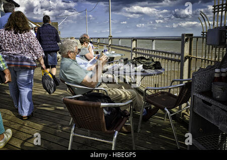 Southwold, England UK. Elderly people enjoying the sunny weather sitting along Southwold pier drinking tea and reading newspapers.  Younger visitors are walking by.  Photograph converted to HDR, High Dynamic Range , for a more dramatic high contrast image. Stock Photo