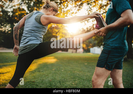 Male coach helping young woman exercising in park. Trainer helping woman in leg stretching workout. Stock Photo