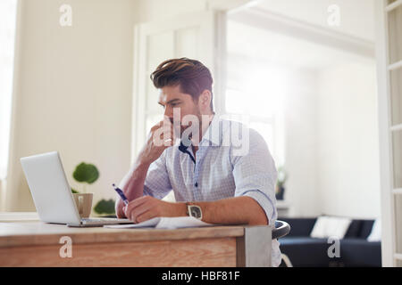 Side view shot of pensive young man sitting at home and working on laptop. Caucasian male working from home office. Stock Photo