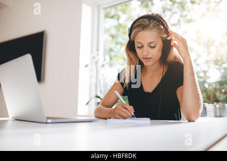 Indoor shot of woman studying in kitchen. Female wearing headphones and writing with laptop on table. Stock Photo