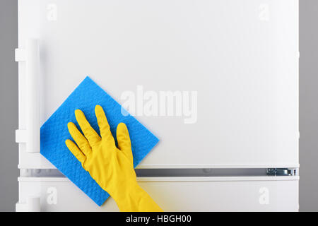 Woman's hand in yellow rubber protective glove cleaning white refrigerator with blue rag on gray background Stock Photo