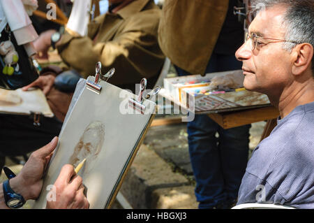 A middle-aged male tourist wearing glasses poses for an artist drawing his portrait at Place du Tertre square in Paris. Stock Photo