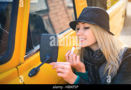 Sweet young woman applies red lipstick looking at the car mirror on a sunny autumn day Stock Photo