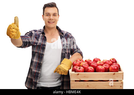 Joyful farmer leaning on a crate filled with apples and giving a thumb up isolated on white background Stock Photo