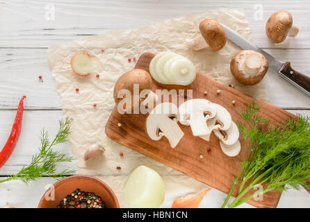 Brown champignon mushrooms on a cutting board. White wooden desk herbs and onions. Stock Photo